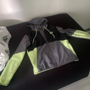 Grå och grön vindjacka från Under Armour vatten tät. - Säljer en snygg vindjacka från Under Armour i grått och grön . Jackan har huva med dragsko, dragkedja framtill och långa ärmar. Perfekt för utomhusaktiviteter och träning. Stilsäker och sportig design!