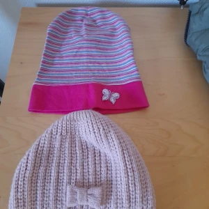 Två mössor med detaljer - En randig mössa i rosa, grått och vitt med rosa kant och en glittrig fjärilsdetalj. Den andra är en beige ribbstickad mössa med en söt rosett framtill.