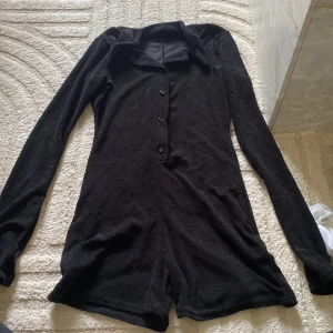 Svart långärmad playsuit från PrettyLittleThing - Svart långärmad playsuit från PrettyLittleThing med krage och knappar framtill. Mjuk och stretchig, perfekt för en trendig och bekväm look. Enkel att styla med sneakers eller boots.