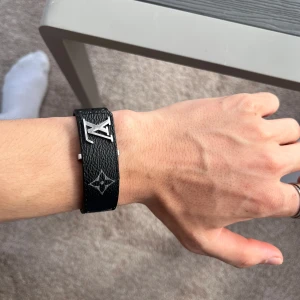 Svart armband från Louis Vuitton - Säljer ett svart armband från Louis Vuitton med klassiskt monogrammönster och ett stort LV-spänne i metall. har en lyxig känsla och är tillverkat i skinn med tydliga vita detaljer.