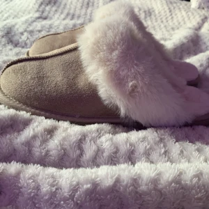 Beige tofflor med fuskpäls - Mysiga beige tofflor med mjuk vit fuskpäls runt öppningen och inuti. Utsidan är i mockaliknande material och sulan är platt. super sköna och mysiga att ha inomhus går att ha ute såklart men inte de dem är till för vad jag vet, aldrig använda, märke vet jag tyvärr inte dom har bara stått i mitt rum hur länge som helst