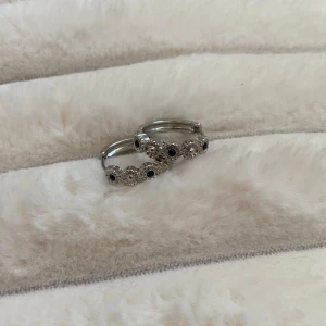 Silverfärgade örhängen med stenar - Säljer ett par runda silverfärgade örhängen med små gnistrande stenar och mörkblå detaljer. Örhängena har en elegant design med dekorativa inslag runt hela ringen. Perfekta för att lyfta din stil.