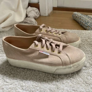 Beige sneakers från Superga med platåsula - Säljer ett par beigea Superga sneakers i mocka med platåsula. Skorna har snörning med guldfärgade öljetter och en klassisk Superga-logga på sidan. Perfekta för dig som gillar en stilren och enkel look.