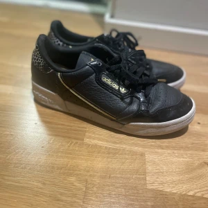 Svarta Adidas sneakers med gulddetaljer - Säljer ett par svarta Adidas sneakers med gulddetaljer och vit sula. Skorna har snörning och ett mönster i form av vita hexagoner på hälen. Adidas-logga i guld på sidan och baksidan. Perfekta för dig som vill ha en stilren men unik sneaker.