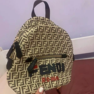 Beige och svart ryggsäck från Fendi - Säljer en beige och svart liten ryggsäck från Fendi med klassiskt FF-mönster och broderad logga framtill. Väskan har dragkedja, justerbara axelband i olika färger och en praktisk framficka. Perfekt accessoar för dig som vill sticka ut!
