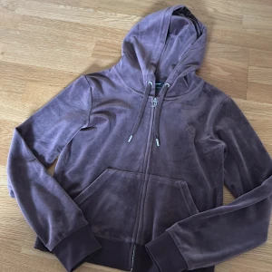 Brun velour hoodie från Juicy Couture - Mysig brun hoodie i velour från Juicy Couture med dragkedja framtill och huva. Tröjan har långa ärmar, fickor och justerbar dragsko vid huvan. Perfekt för en avslappnad och trendig look.