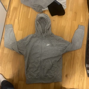 Grå hoodie med dragkedja från Nike - Säljer en grå hoodie från Nike med dragkedja och huva. Jackan har två fickor framtill och en liten vit Nike-logga på bröstet. Perfekt för en avslappnad stil och enkel att matcha med det mesta. Har rak passform i storlek S, men passar även strlk M