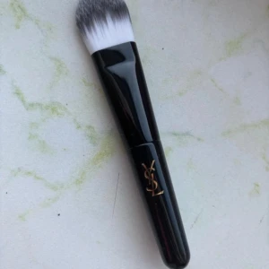 Mini sminkborste från Yves Saint Laurent - Snygg sminkborste från Yves Saint Laurent med svart handtag och vita till grå borsthår. Perfekt för applicering av foundation eller concealer. Handtaget har en elegant guldig YSL-logga. Mini-storlek. Oanvänd 