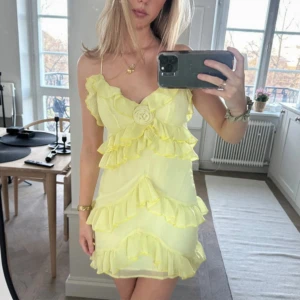 Gul volangklänning med smala axelband - Söt ljusgul kortklänning med flera volanglager och smala axelband. Klänningen har en dekorativ blomdetalj framtill och är tillverkad i ett lätt, luftigt material. Perfekt för varma dagar och somriga tillfällen. Bild från tidigare ägare då den tyvärr inte passa mig