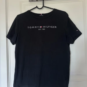 Svart t-shirt från Tommy Hilfiger - Säljer en svart t-shirt från Tommy Hilfiger med klassisk logga och text på bröstet. T-shirten är i mjuk bomull och har rund halsringning samt korta ärmar. Perfekt för en stilren och enkel look.