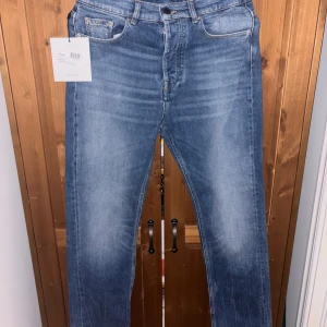Blå jeansbyxor från J.Lindeberg - Snygga blå jeansbyxor från J.Lindeberg i vintage wash med klassisk femficksdesign och läderpatch bak. Jeansen är tillverkade i ekologisk bomull och har en rak passform med normal midja. Perfekta för en stilren vardagslook. Köpta för 1600, oanvända.