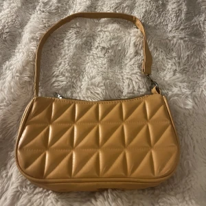 Beige quiltad axelväska - Säljer en beige axelväska med quiltat mönster och dragkedja upptill. Väskan har en enkel, stilren design och är tillverkad i syntetmaterial. Perfekt för dig som vill ha en trendig accessoar med lite retrovibbar.