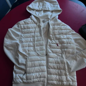 Vit hybridjacka med huva från Moncler - Säljer en vit hybridjacka från Moncler med quiltad front och ärmar i sweatshirtmaterial. Jackan har huva med dragsko, dragkedja framtill och två fickor med dragkedja. På ena ärmen finns en ficka med lock och Moncler-logga.