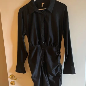 Svart skjortklänning från NLY Trend - Svart skjortklänning från NLY Trend med lång ärm och knappar framtill i satinmaterial. Klänningen har krage och draperad detalj i midjan som ger en snygg form och knappt använd🖤