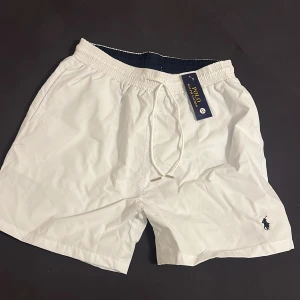 Vita badshorts - Säljer ett par fräscha vita badshorts från Polo Ralph Lauren med klassisk broderad logga på benet. Shortsen har snörning i midjan, innerbyxa i mesh och en bakficka. Perfekta för sommaren! Liten i storlek!