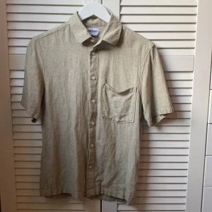 Beige linneskjorta från Weekday - Säljer en stilren beige skjorta i linne från Weekday. Skjortan har korta ärmar, klassisk krage, bröstficka och knäppning framtill. Perfekt för varma dagar och enkel att matcha med olika outfits.