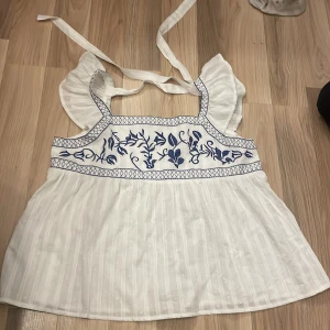 Vit blus med blå broderi och volangärmar, storlek L - Superfin vit blus med blått broderat blommönster över bysten och volangärmar. Blusen har knytband på axlarna och är tillverkad i ett lätt, luftigt tyg med diskreta ränder. Perfekt för varma dagar!