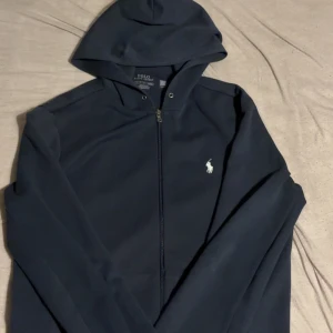 Mörkblå hoodie från Polo Ralph Lauren - Mörkblå hoodie med dragkedja från Polo Ralph Lauren. Klassisk design med vit broderad logga på bröstet och huva. Perfekt för en avslappnad stil.