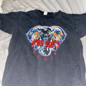 Svart Kenzo t-shirt med elefanttryck - Cool svart t-shirt från Kenzo med stort färgglatt elefanttryck och röd Kenzo Paris-text på bröstet. Tillverkad i 100% bomull och har en klassisk passform med korta ärmar. Perfekt för dig som gillar statement-plagg! Liten i storlek 