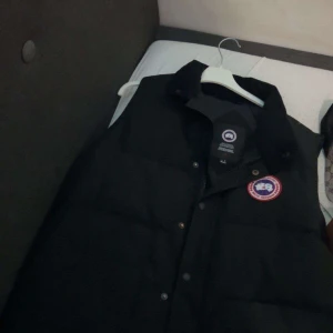 Svart dunväst från Canada Goose - Säljer en svart dunväst från Canada Goose med klassisk logotyp på bröstet. Västen har knappar framtill och är perfekt för kyligare dagar. Stilsäker och tidlös design som passar till många outfits. Pris kan diskuteras vid snabb affär!