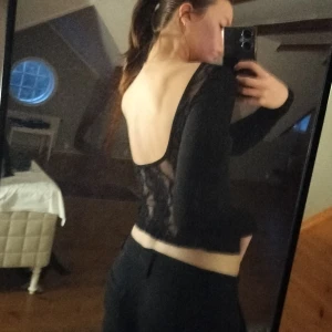 Svart långärmad topp med spetsdetaljer - Säljer en svart långärmad topp med transparenta spetsdetaljer på sidorna och öppen rygg. Toppen är tight och har en stilren, elegant look med blommig spets som ger en unik touch. Aldrig använd bara testad 