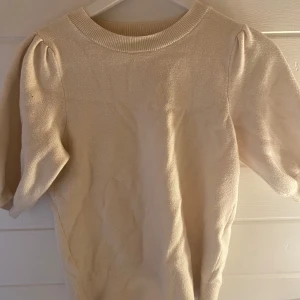 Beige stickad kortärmad tröja - Säljer en söt beige stickad tröja med korta puffärmar och rund halsringning från lager157. Perfekt att styla till både jeans och kjol för en trendig look. Tröjan har en mjuk känsla och ribbad kant vid halsen.