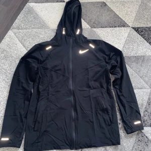 Nike Miler - Svart vindjacka från Nike med huva och dragkedja framtill. Jackan har reflekterande detaljer och två fickor med dragkedja. Perfekt för utomhusaktiviteter och har en sportig look.