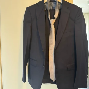 Mörkblå kostym med kavaj och byxor - Säljer en klassisk mörkblå kostym med matchande kavaj och byxor. Kavajen har två knappar framtill, fickor med lock och en slät passform. Byxorna är raka och stilrena. Storlek på byxor 30x30, storlek på kavaj 44 regular. 