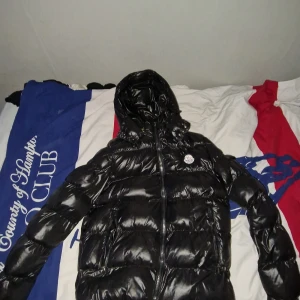 Svart pufferjacka från Moncler - Säljer en svart glansig pufferjacka från Moncler med huva och dragkedja. Jackan har en klassisk Moncler-logga på bröstet och två fickor framtill. Perfekt för kyliga dagar när du vill hålla värmen och ändå se stilren ut.