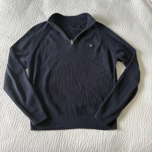 Mörkblå stickad tröja med dragkedja från GANT - Säljer en mörkblå stickad tröja från GANT med halv dragkedja och broderad logga på bröstet. Tröjan har lång ärm, ribbad struktur och hög krage. Perfekt för en stilren och klassisk look. Finns inga defekter. Bara att höra av sig vid minsta fundering!