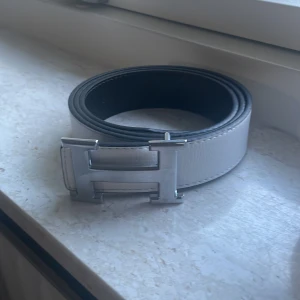 Vitt skärp med silverfärgat H-spänne - Snyggt vitt bälte med ett stort, silverfärgat H-format spänne. Bältet har en stilren design och passar perfekt till jeans eller kostymbyxor. Perfekt accessoar för att lyfta din outfit.
