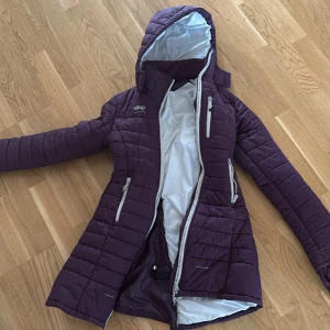 Lila quiltad jacka med huva från Uhip - Säljer en lila quiltad jacka från Uhip med huva och dragkedja. Jackan har långa ärmar, flera fickor med dragkedja och en snygg passform. Perfekt för kyliga dagar och har en längre modell baktill.