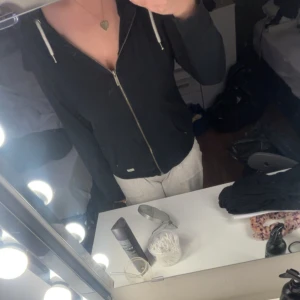 Svart hoodie med dragkedja - Svart hoodie med dragkedja och huva. Modellen har långa ärmar och vita snören vid huvan. Perfekt för en avslappnad stil.