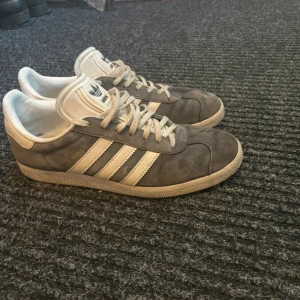 Grå Adidas Gazelle sneakers - Säljer ett par klassiska Adidas Gazelle sneakers i grå mocka med vita detaljer och vita ränder på sidorna. Skorna har snörning och platt sula, samt Adidas-logga på plösen och hälen. Perfekta för en stilren vardagslook. Nypris 1200kr mitt pris 500kr priset är dock inte hugget i sten. 