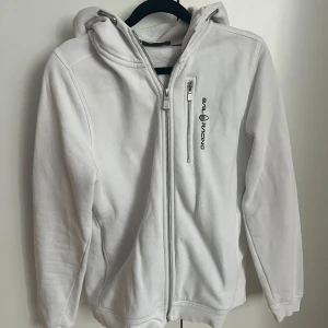 Vit hoodiejacka från Sail Racing - Vit hoodiejacka från Sail Racing med dragkedja framtill och en extra dragkedjeficka på bröstet. Jackan har huva och logga på bröstet samt baktill på huvan. Perfekt för en sportig och avslappnad stil.