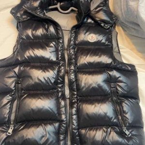 Svart dunväst från Moncler - Snygg svart dunväst från Moncler med glansig finish och två dragkedjefickor framtill. Västen har hög krage och Moncler-logga på bröstet. Insidan är ljusgrå med klassisk Moncler-etikett och serietidningsdetalj.möts i Norrköping 