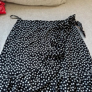 Svart omlottkjol med blommigt - Supersöt svart omlottkjol från SHEIN med vitt blommigt mönster och volang. Kjolen knyts i midjan🖤