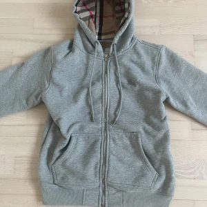 Burberry kofta - Säljer en grå hoodie/kofta från Burberry med klassiskt rutigt foder i huvan. Jackan har dragkedja framtill, två fickor och ribbade muddar. Perfekt för en avslappnad och stilren look.