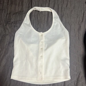 Vit ribbad halterneck-linnetopp från Dazy - Säljer en vit ribbad halterneck-linnetopp från Dazy med knappar framtill. Toppen har en croppad passform och är ärmlös, perfekt för varma dagar. Enkel och stilren design som passar till mycket.