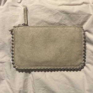 Beige kuvertväska med silverdetaljer från Zara - Säljer en beige kuvertväska från Zara med dragkedja och silvriga pärldetaljer runt kanterna. Väskan har flera fack inuti och är perfekt att bära i handen. Enkel och stilren design som passar till många outfits.