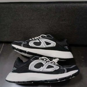 Svarta och vita sneakers från Dior - Säljer ett par svarta och vita sneakers från Dior med tydlig logga på plösen och insidan. Skorna har vit sula, svarta meshpaneler och vita detaljer i syntetmaterial. Klassisk snörning och sportig design.  Helt nya ‼️med kvitto 