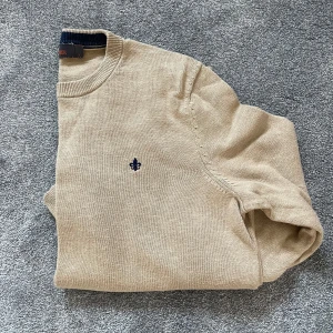 Beige stickad tröja från Morris - Säljer en klassisk beige stickad tröja från Morris med rund halsringning och diskret broderad logga på bröstet. Tröjan har långa ärmar och ribbade muddar vid hals, ärmslut och nederkant. Perfekt för lager-på-lager under kyliga dagar. Pris kan diskuteras vid snabb affär. 