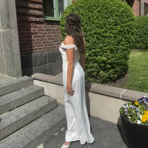 Vit långklänning med offshoulder och oneshoulder-detalj - Elegant vit långklänning med en unik kombination av offshoulder och oneshoulder-design. Klänningen har en figurnära passform och är tillverkad i ett glansigt material som ger en lyxig känsla. Perfekt för speciella tillfällen. Från Adoore, använd en gång!