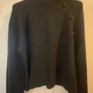 Svart stickad tröja med knappar - Mysig svart stickad tröja med långa ärmar och dekorativa knappar längs axeln. Perfekt för kyligare dagar och enkel att matcha med olika outfits.