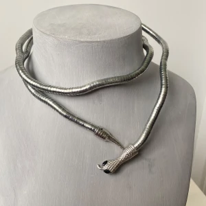 Silvrigt ormformat halsband - Unikt halsband i silverfärgad metall, format som en orm med detaljerat ormansikte och svans i ändarna. Halsbandet har en böjlig, segmenterad design som ger en cool och edgy look. Perfekt för dig som vill sticka ut med accessoarer.