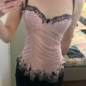 Ljusrosa korsettopp med spets - Supersöt ljusrosa korsettopp från Ann Christine med mörklila spetsdetaljer, små rosetter och volang nertill. Toppen har reglerbara axelband. Köpt på vinted men använd 1 gång av mig