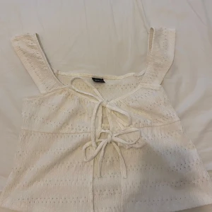 Vit linnetopp med knytning från Gina Tricot - Säljer en söt vit linnetopp från Gina Tricot med breda axelband och dekorativa knytningar framtill. Toppen har ett diskret hålmönster i tyget och är perfekt för varma dagar.