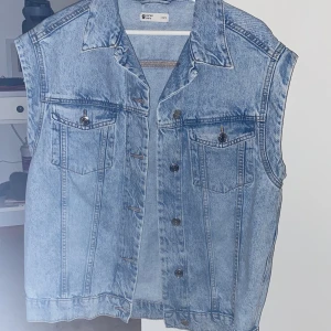 Ljusblå jeansväst från Gina Tricot - Säljer en klassisk ljusblå jeansväst från Gina Tricot med knappar framtill och två bröstfickor med lock. Västen har en rak passform och är ärmlös, perfekt att styla över en t-shirt eller hoodie.