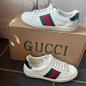 Gucci ace  sneakers med gröna och röda ränder - Säljer ett par vita Gucci ace sneakers med klassiska gröna och röda ränder på sidan och metallicgrön häl. Skorna har vit snörning och platt sula. Kommer med originalkartong, extra skosnören och dustbag.