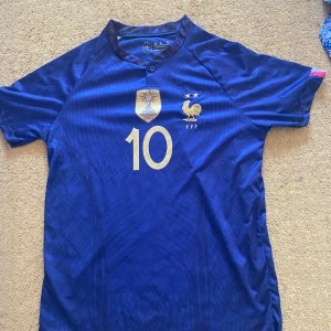 Frankrike Mbappé 10 matchtröja - Säljer en blå fransk landslagströja med guldiga detaljer, nummer 10 och Mbappé på ryggen. Tröjan har korta ärmar, broderad tupp-logga och VM-märke på bröstet. Perfekt för fotbollsfans som vill stötta Frankrike!